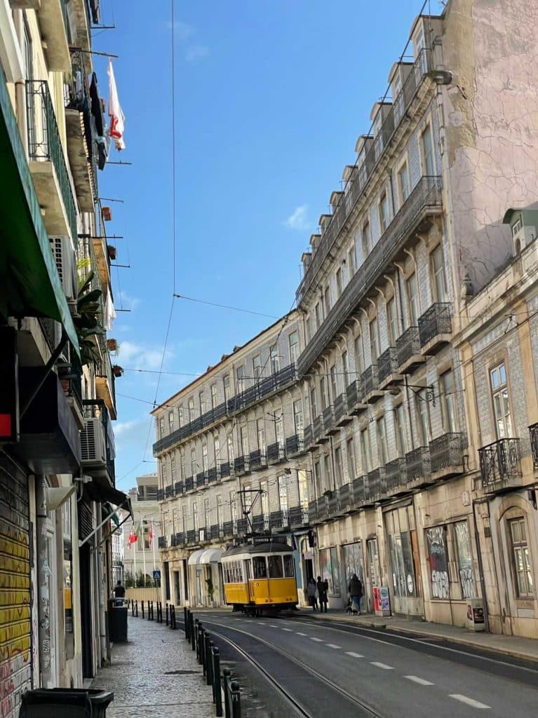 Een gele tram rijdt door een smalle straat met oude, sierlijke gebouwen in Lissabon, Portugal - een must voor bezoekers terwijl voetgangers over het trottoir wandelen onder een strakblauwe hemel.