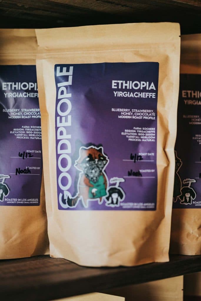 Een zak GOODPEOPLE Ethiopië Yirgacheffe koffie met een paars label met een cartoon wasbeer. Op het etiket staat dat deze koffielanden werd bezocht en geproefd, met proefnotities van bosbes, aardbei, honing, chocolade en de branddatum.