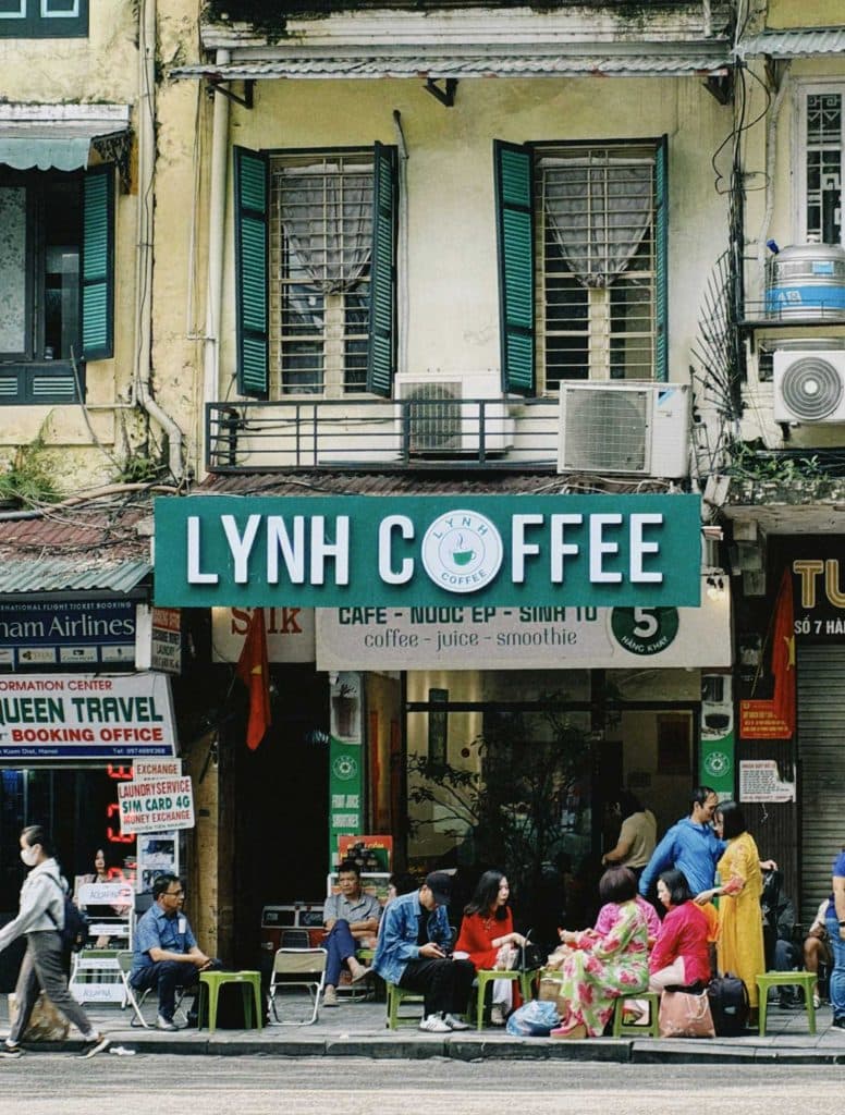 Mensen zitten op kleine krukjes buiten "Lynh Coffee", een drukke plek op de begane grond van een oud geel gebouw met groene luiken. Velen hebben deze lokale favoriet bezocht om de charme te ervaren waar koffielanden beroemd om zijn.