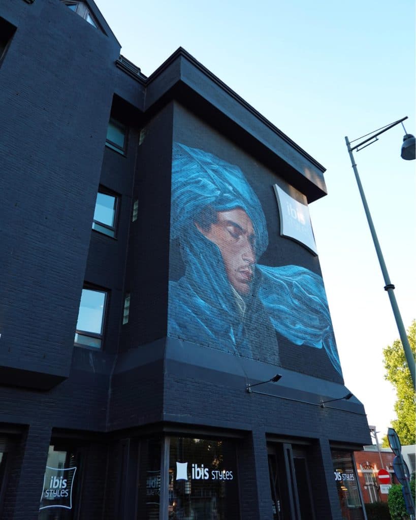 Een muurschildering op de buitenmuur van een donker gebouw in Hasselt toont een persoon met gesloten ogen, gedrapeerd in een vloeiende blauwe stof. Het gebouw herbergt een Ibis Styles hotel, zoals de bewegwijzering aangeeft, en wordt verlicht door daglicht.