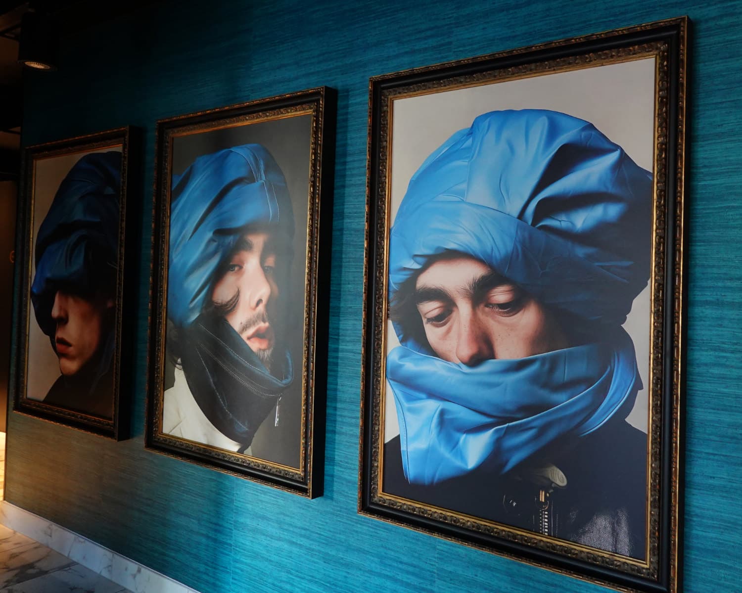 Drie ingelijste portretten op een groenblauwe muur tonen mensen met blauwe gewatteerde kappen, die doen denken aan Antarctica-uitrusting, om hun hoofd gewikkeld. De belichting benadrukt de texturen van de stof en de contemplatieve uitdrukkingen van de geportretteerden.