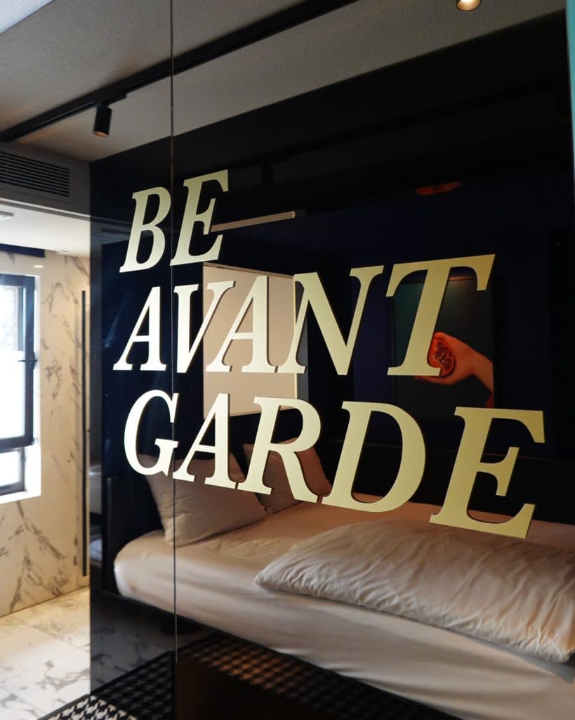 Grote beige letters met de tekst "BE AVANT GARDE" zijn gedrukt op een reflecterende glazen muur in een moderne, stijlvolle kamer met marmeren accenten en een geruite vloer - een vleugje Hasseltse flair ver van Antarctica.