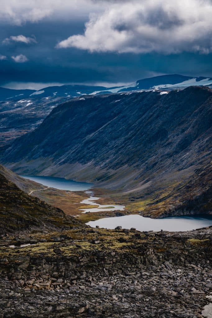 Een dramatisch berglandschap met donkere, steile kliffen, verspreide sneeuwvlokken en een kronkelend fjord onder een bewolkte, stemmige lucht. De rotsachtige voorgrond en gelaagde heuvels roepen de wilde schoonheid op die Noorwegen Nomads omarmt.