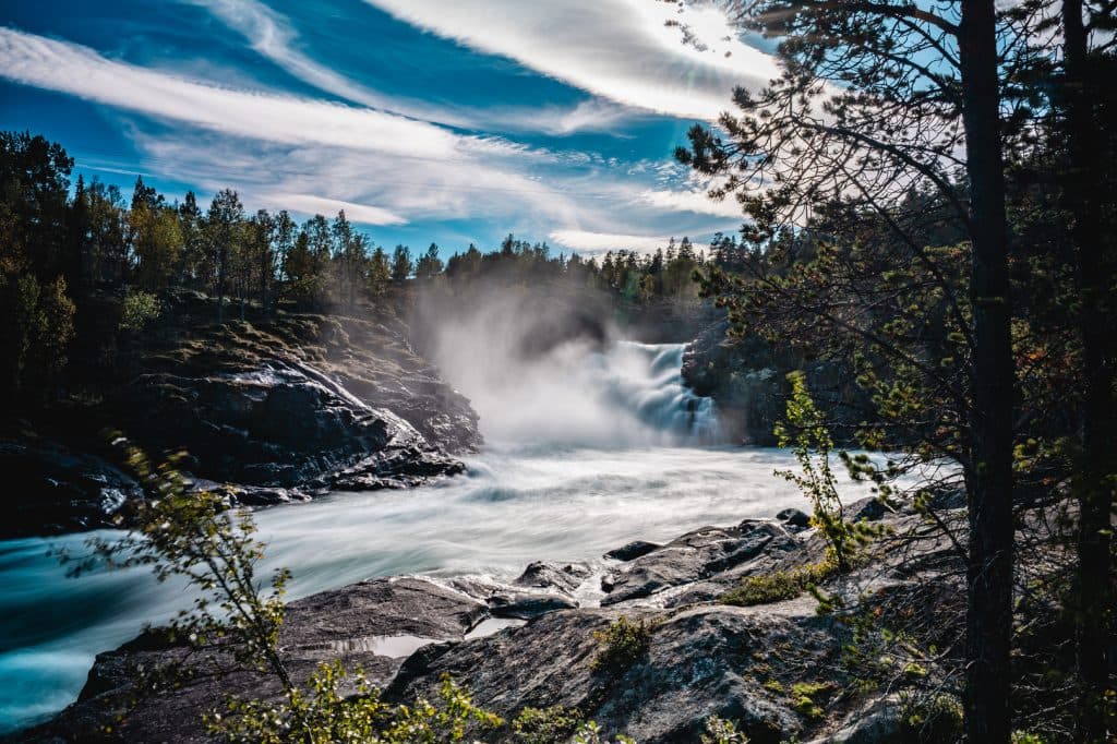 Een krachtige waterval stort zich uit over rotsachtig terrein in fjordenland, omringd door pijnbomen en mist. Dramatische, gestreepte wolken vullen een helderblauwe hemel terwijl het zonlicht van de middernachtzon er doorheen schijnt en dit Noorwegen Nomads avontuur verlicht.