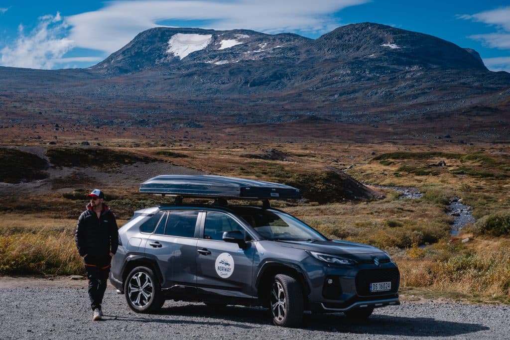 Een persoon in outdoorkleding staat naast een grijze SUV met dakkoffer, geparkeerd op een grindweg onder de middernachtzon, met fjorden en bergen die zich uitstrekken aan de horizon-Noorwegen Nomaden die de wilde schoonheid verkennen.