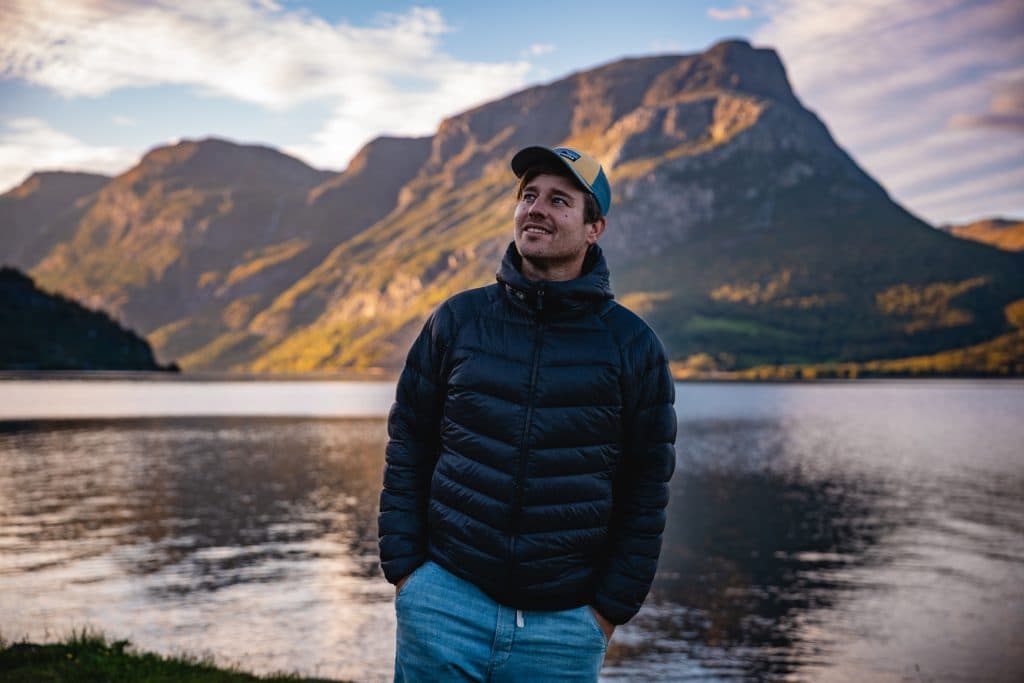 Een man in een zwarte pufferjas en een blauwe pet staat met zijn handen in zijn zakken te glimlachen bij een rustig fjordenmeer, terwijl op de achtergrond hoge bergen en een bewolkte lucht oprijzen - typerend voor de avonturen van Noorwegen Nomads.