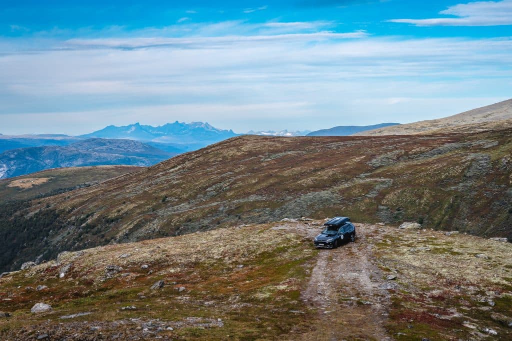 Een zwarte SUV staat geparkeerd op een rotsachtig, onverhard pad boven op een bergrug met verre bergtoppen onder de middernachtzon. Het landschap, dat doet denken aan de fjorden, bestaat uit grashellingen, rotsen en weidse uitzichten die geschikt zijn voor Noorwegen Nomaden.