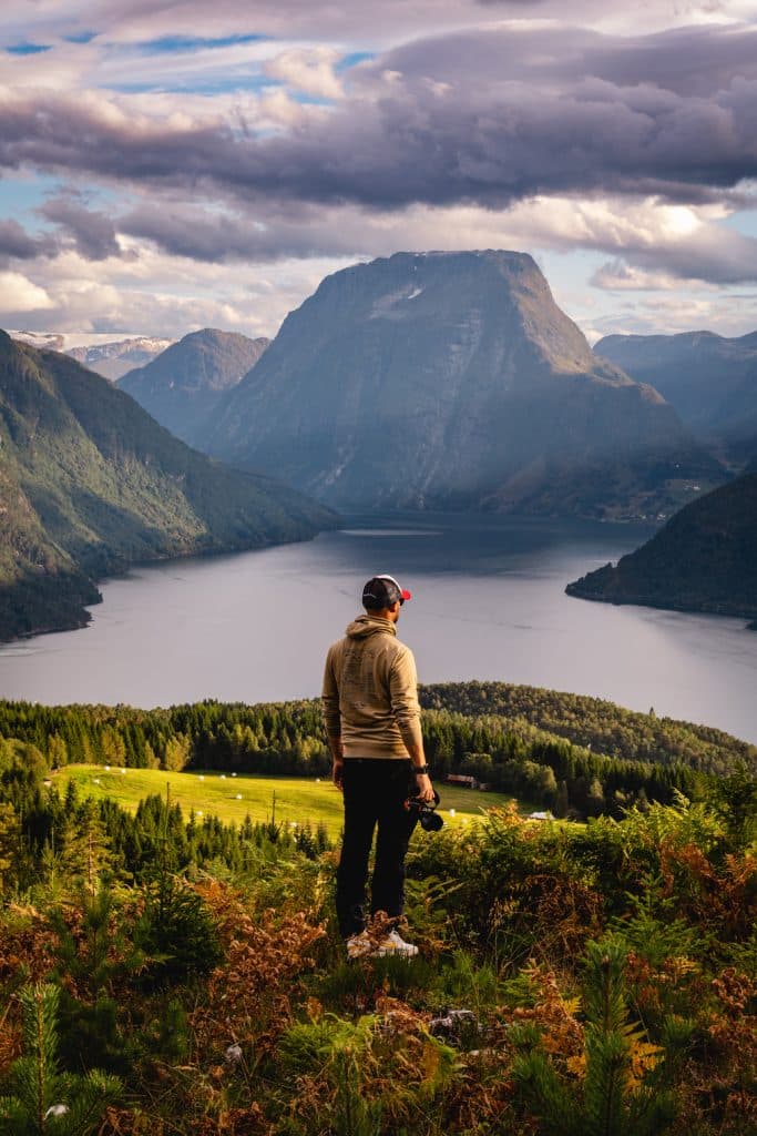 Een persoon staat op een met gras begroeide heuvel en kijkt uit over de Noorwegen Nomads fjorden, omringd door beboste bergen en dramatische wolken, met een grote, prominente piek op de achtergrond onder de gloed van de middernachtzon.