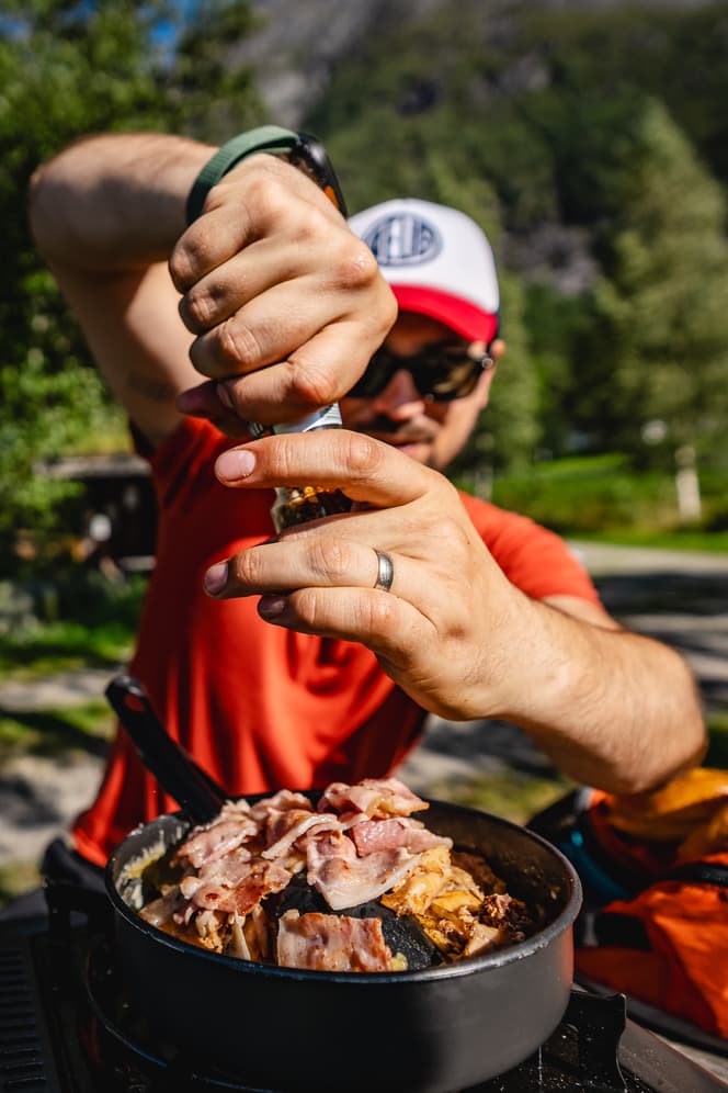 Een persoon met een zonnebril op en een pet op maalt buiten peper op een pan gevuld met spek en ander voedsel, met bomen en bergen zichtbaar op de achtergrond, waarmee de geest van Noorwegen Nomads onder de majestueuze fjorden wordt vastgelegd.