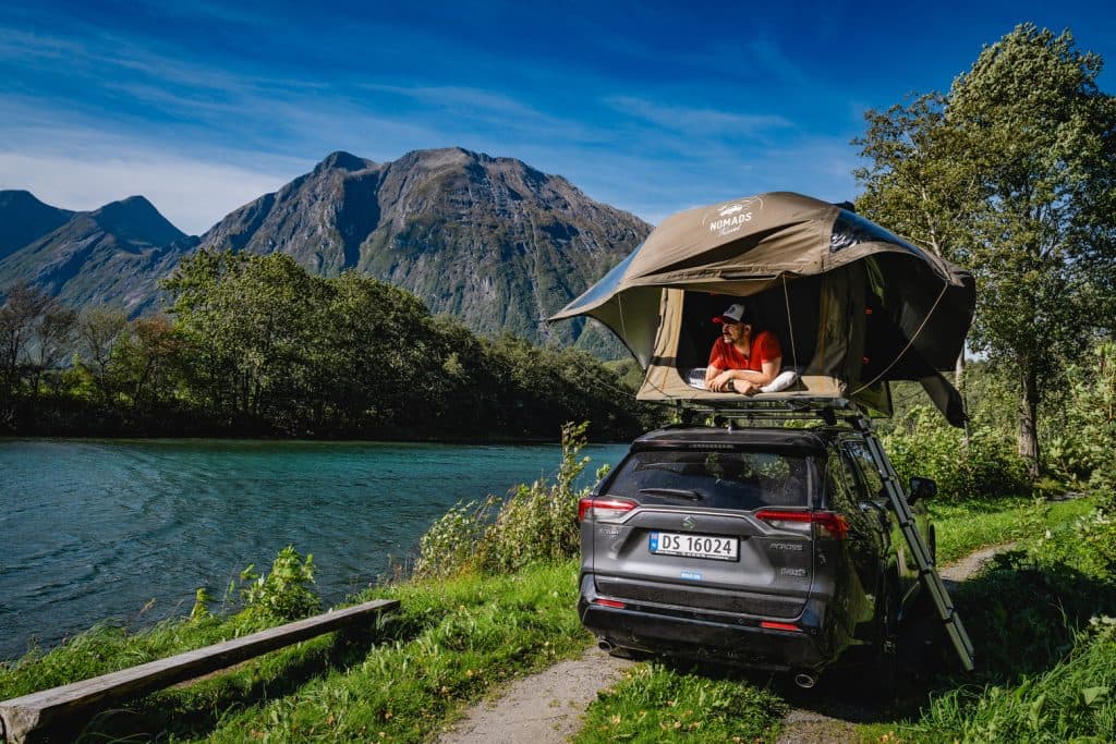 Een persoon in een rood shirt zit in een daktent op een zwarte SUV bij een rivier, omringd door groene bomen en hoge bergen, onder een strakblauwe hemel - een ideaal tafereel voor Noorwegen Nomads die de fjorden verkennen onder de middernachtzon.
