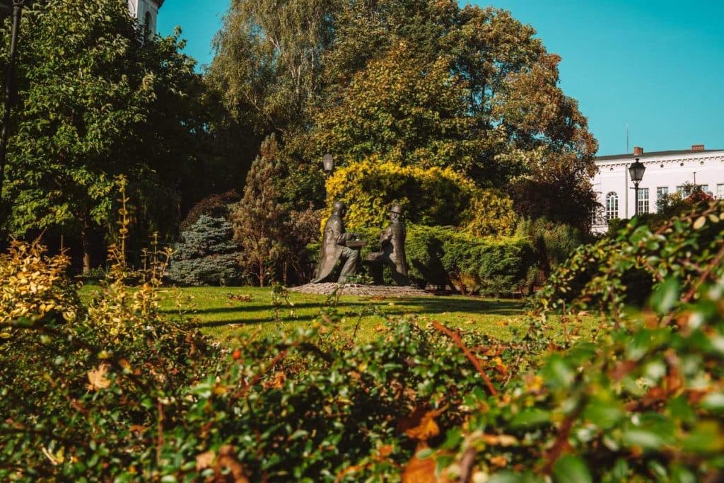 Een weelderig park met dicht groen en lommerrijke bomen omringt een beeld van drie mensen die aan een tafel zitten. Fel zonlicht accentueert het gebladerte en op de achtergrond is een wit gebouw zichtbaar - een rustige plek voor Opole reizen.