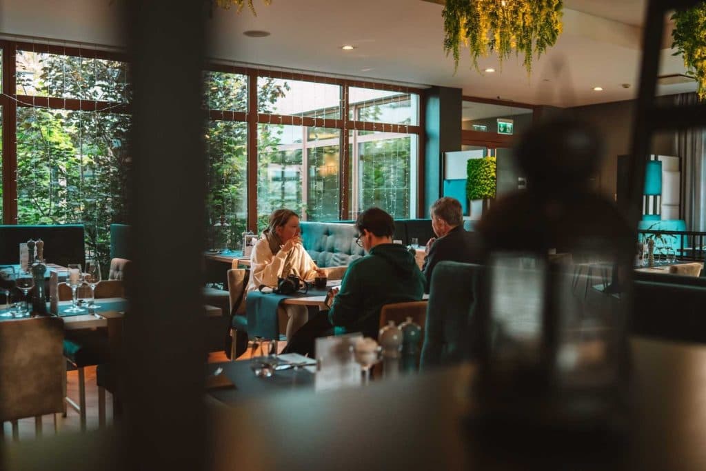 Drie mensen zitten aan een tafel in een modern, zonovergoten restaurant in Opole, met grote ramen, weelderig groen buiten en hangende planten binnen, en genieten van een levendig gesprek - een ideaal moment voor Opole reizen liefhebbers.