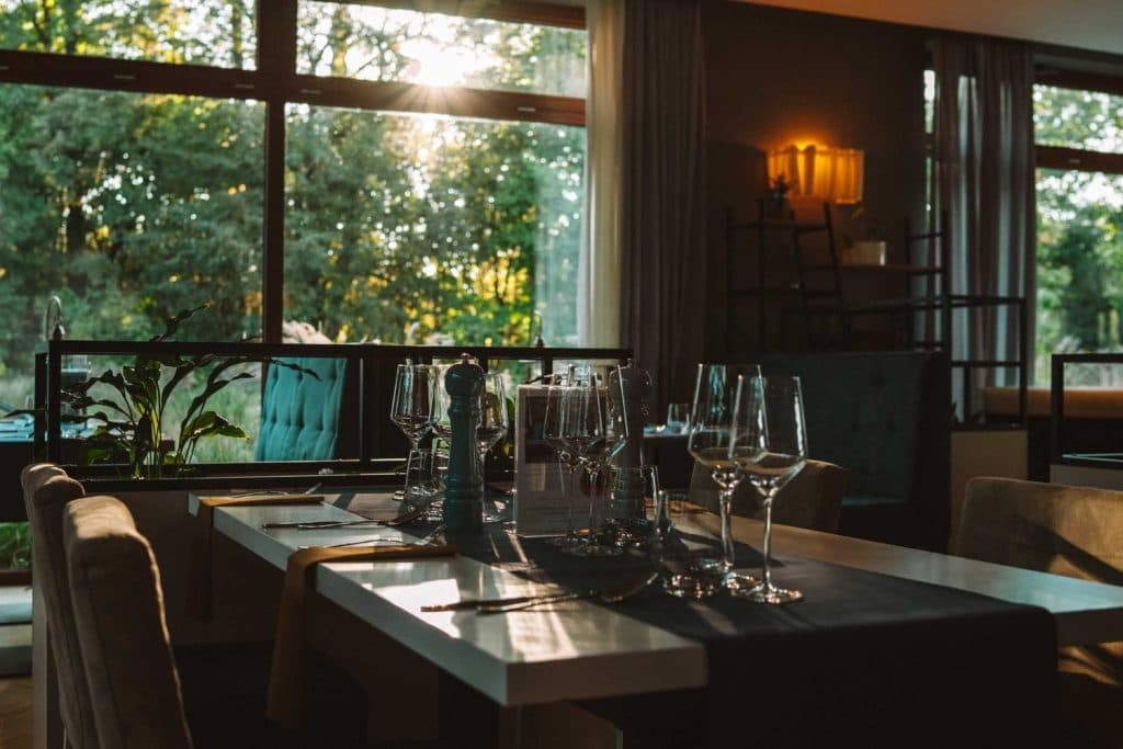 Een gedekte eettafel met wijnglazen, bestek en een pepermolen in een modern restaurant met grote ramen - het zonlicht stroomt naar binnen met uitzicht op de groene bomen buiten - een perfecte plek om te genieten van Opole reizen.