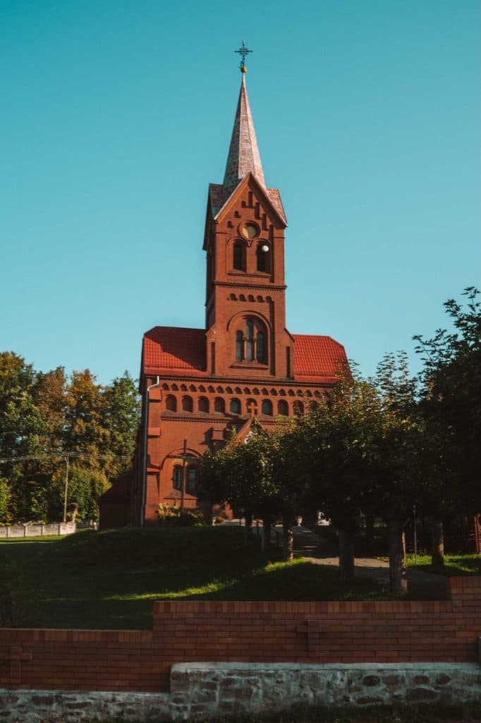 Een hoge kerk van rode baksteen met een spitse toren en gewelfde ramen staat tussen groene bomen onder een helderblauwe hemel, waardoor het een bezienswaardigheid is voor iedereen die van Opole reizen geniet.
