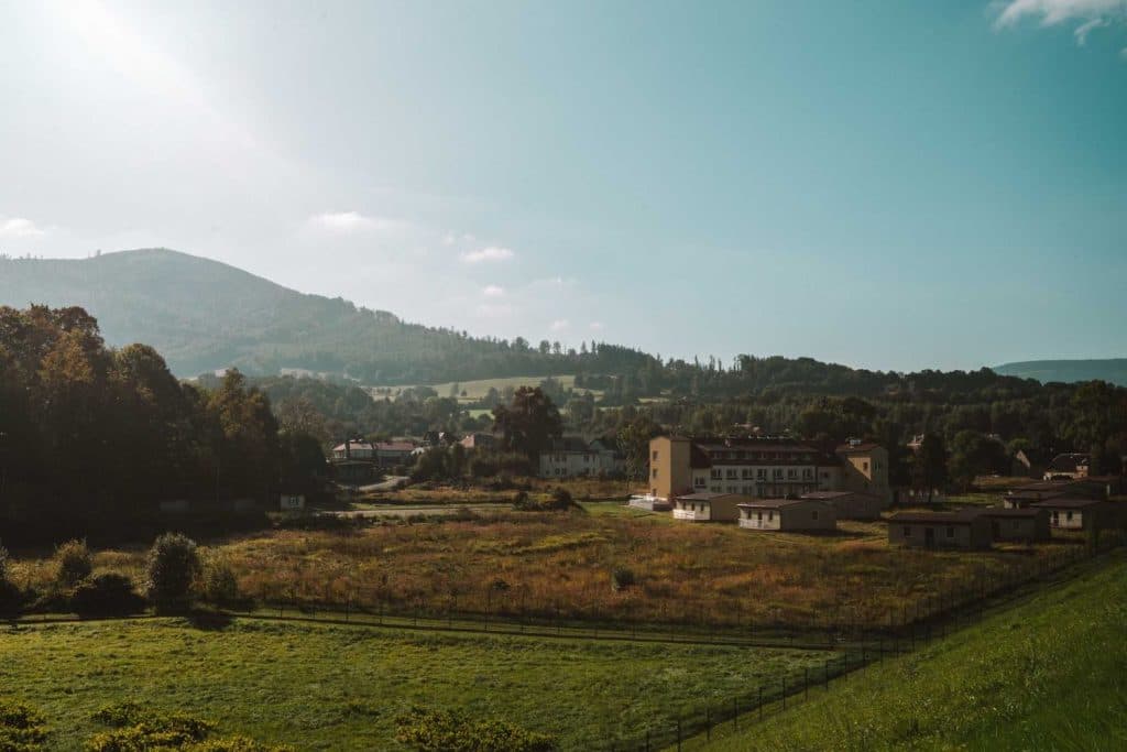 Een landelijk landschap met grasvelden, verspreide bomen en een paar gebouwen op de voorgrond - perfect voor Opole reizen - tegen glooiende heuvels en een helderblauwe lucht. Zonlicht werpt lange schaduwen over de scène.
