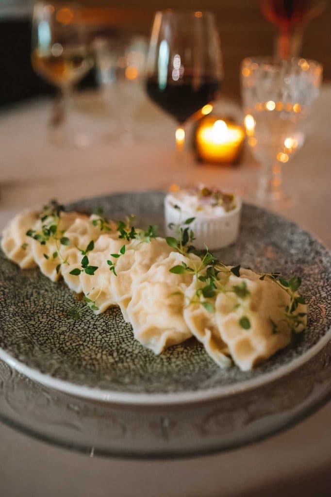 Een bord knoedels gegarneerd met verse kruiden ligt op een schaal met een patroon, perfect voor wie geniet van Opole reizen, vergezeld van een kom witte dipsaus. Wijnglazen en een brandende kaars op de achtergrond maken de gezellige, elegante dinersfeer compleet.