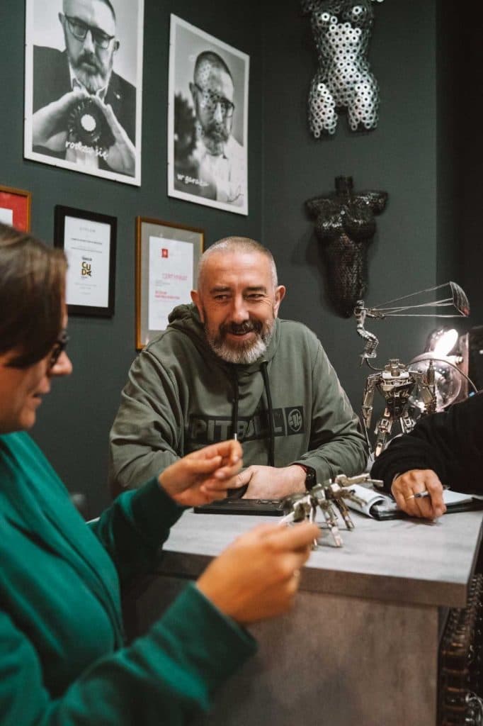 Een bebaarde man met een groene capuchon glimlacht terwijl hij praat met een vrouw met een bril aan een balie, met kunst en ingelijste foto's op de donkere muur achter hen. Ze houdt een klein voorwerp vast, wat duidt op gedeelde herinneringen aan Opole reizen tussen de metalen sculpturen op het bureau.