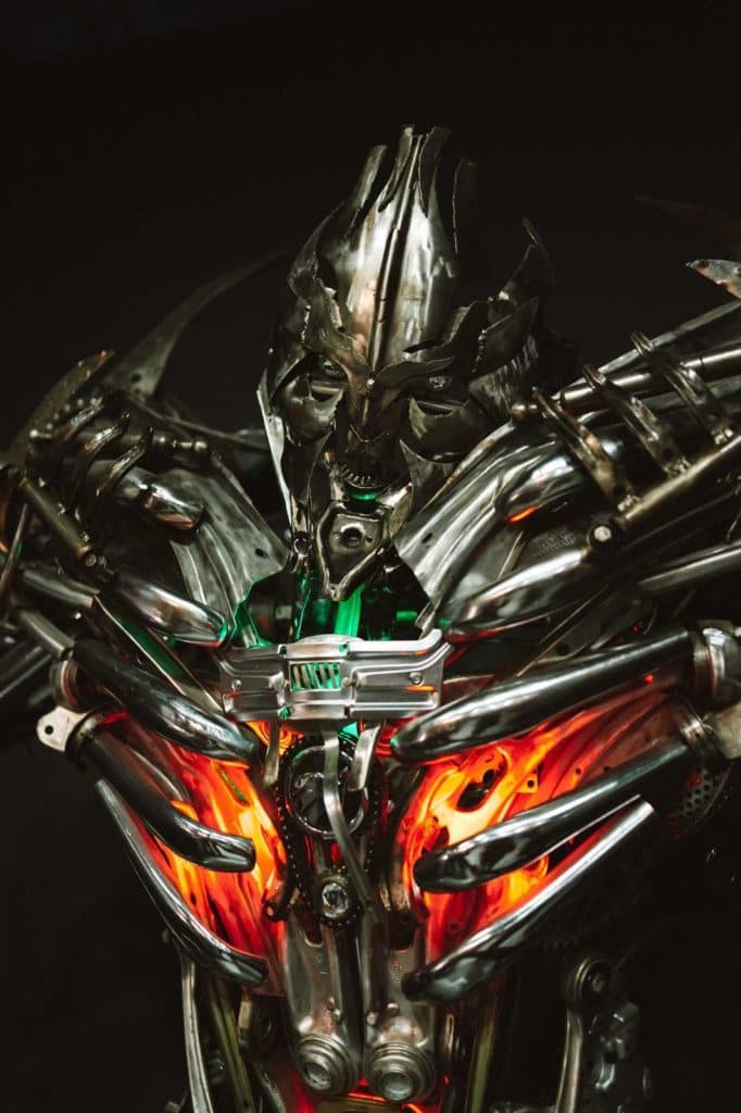 Een dreigende robotfiguur gemaakt van metalen onderdelen met oplichtende oranje en groene lampjes in zijn borst, gedetailleerd met scherpe, hoekige gelaatstrekken, wordt verlicht tegen een donkere achtergrond als een futuristische bewaker tijdens een Opole reizen avontuur.