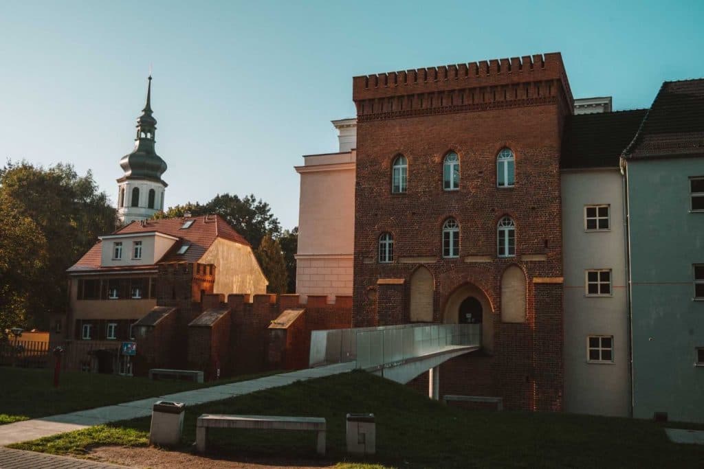 Een bakstenen gebouw in middeleeuwse stijl met boogramen en een moderne glazen brug ligt naast pastelkleurige huizen en een kerk met een groene, gelaagde torenspits onder een helderblauwe hemel - perfect voor wie op zoek is naar unieke Opole reizen ervaringen.
