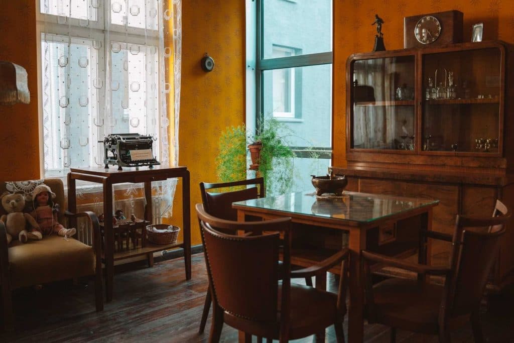Een kamer in vintage stijl met geel behang, houten meubels, een kast met glazen voorkant, een tafel met stoelen en een typemachine bij het raam roept de charme van Opole reizen op. Zonlicht valt door de gordijnen op een bank met teddyberen en een pop.