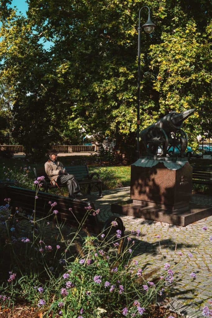 Een man zit op een bankje te lezen in een zonnig park in Opole, op reis door de geschiedenis naast een groot standbeeld van een oud kanon, omringd door groene bomen en paarse bloemen.