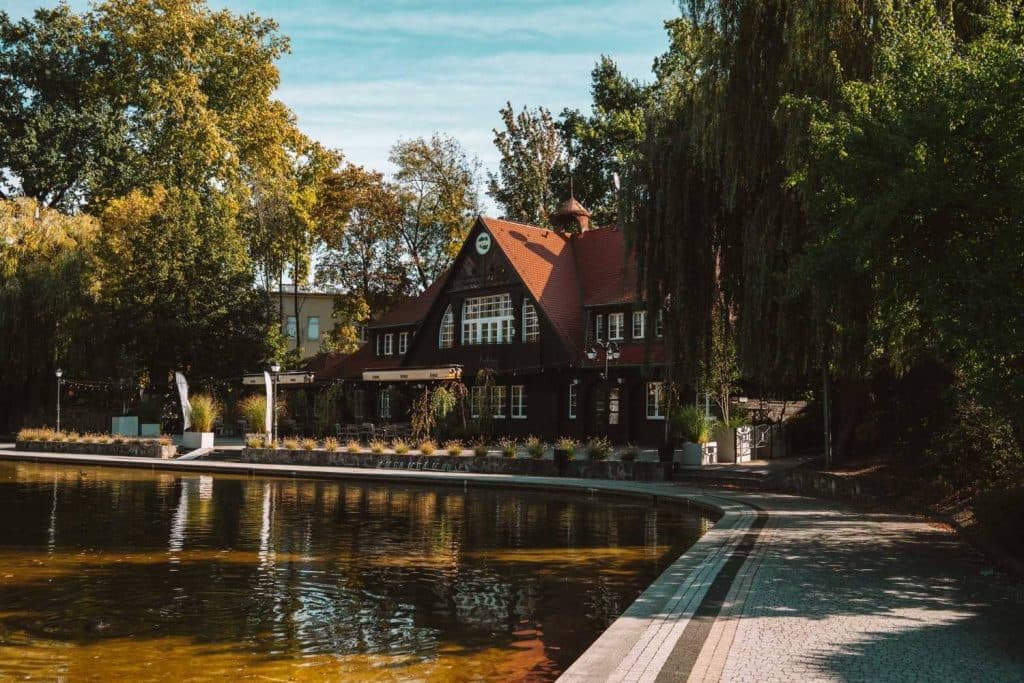 Een charmant, donker houten gebouw aan de oever van het meer met een rood dak ligt naast een wandelpad met bomen en rustig water onder een blauwe hemel, waardoor het een idyllische plek is voor Opole reizen te midden van weelderig groen gebladerte.