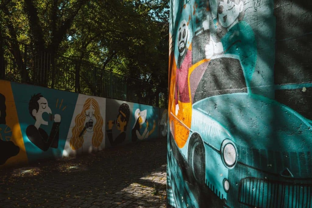 Een muurschildering op een buitenmuur in Opole reizen toont mensen in cartoonstijl met verschillende uitdrukkingen, waaronder een bebaarde man die een auto bestuurt en anderen die zingen, drinken of praten, terwijl bomen en zonlicht schaduwen werpen op het tafereel.