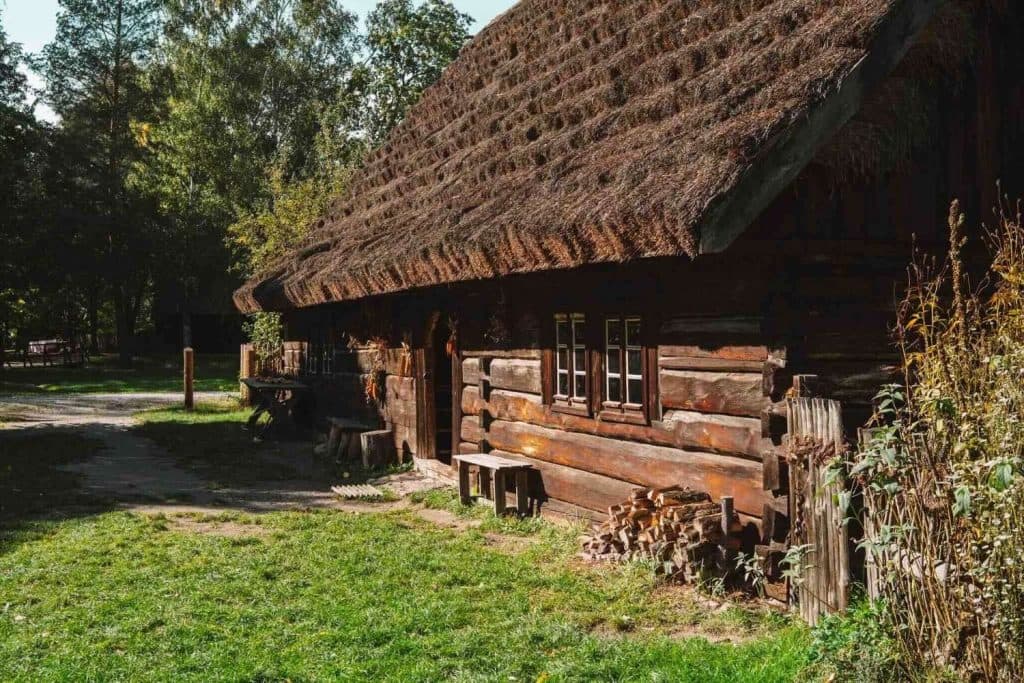 Een rustieke houten hut met rieten dak ligt tussen de bomen en het gras en nodigt bezoekers van Opole reizen uit om tot rust te komen. Houtblokken liggen opgestapeld bij de muur en houten banken en een tafel liggen in het zonlicht, waardoor een vredig landelijk tafereel ontstaat.