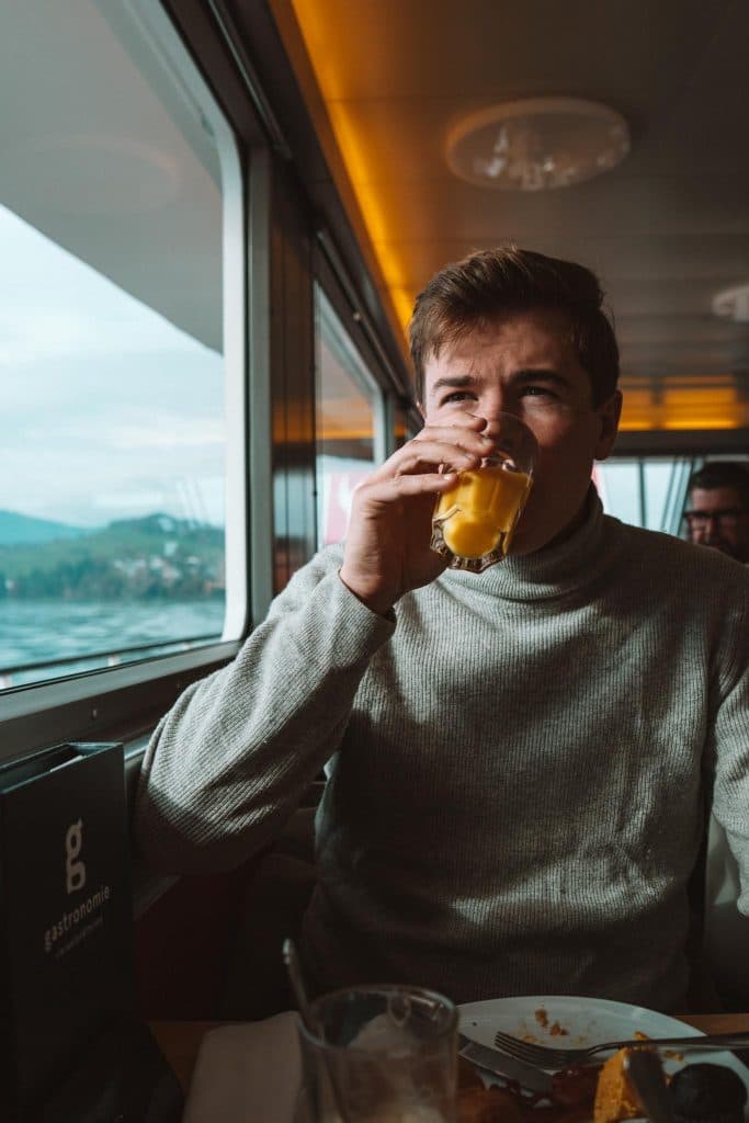 Een man in een grijze coltrui zit aan het raam van een trein of boot en drinkt sinaasappelsap. Zonlicht stroomt naar binnen en voor hem staat eten op tafel. Een andere persoon is gedeeltelijk zichtbaar op de achtergrond.