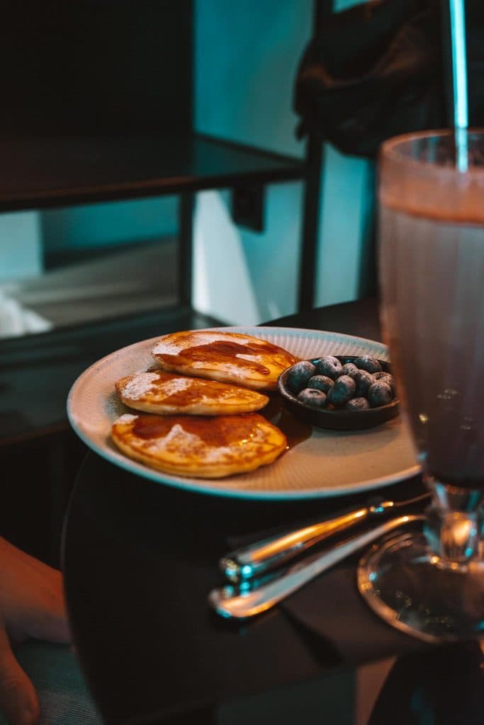 Een bord met kleine pannenkoekjes met stroop en poedersuiker, verse bosbessen en een glas chocolademilkshake op een donkere tafel met een vork en mes naast het bord.