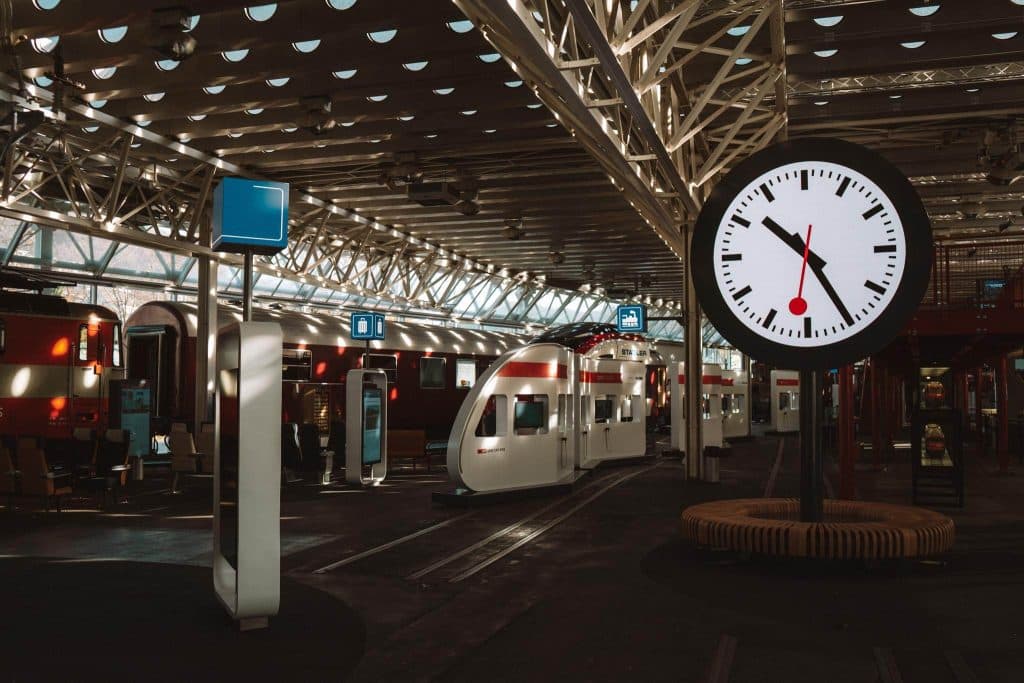Een modern overdekt treinstation met verschillende treinen, digitale schermen en een grote ronde klok die de tijd 2:10 aangeeft. Zonlicht stroomt door de ramen en het geometrische metalen geraamte van het plafond.