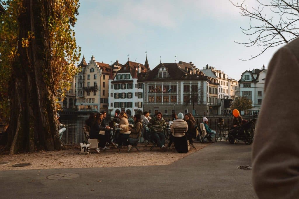 Een groep mensen zit aan een tafel buiten bij een rivier, te kletsen en te ontspannen onder herfstbomen, met historische Europese gebouwen op de achtergrond. Een kind met een oranje ballon en kinderwagens zijn ook zichtbaar.