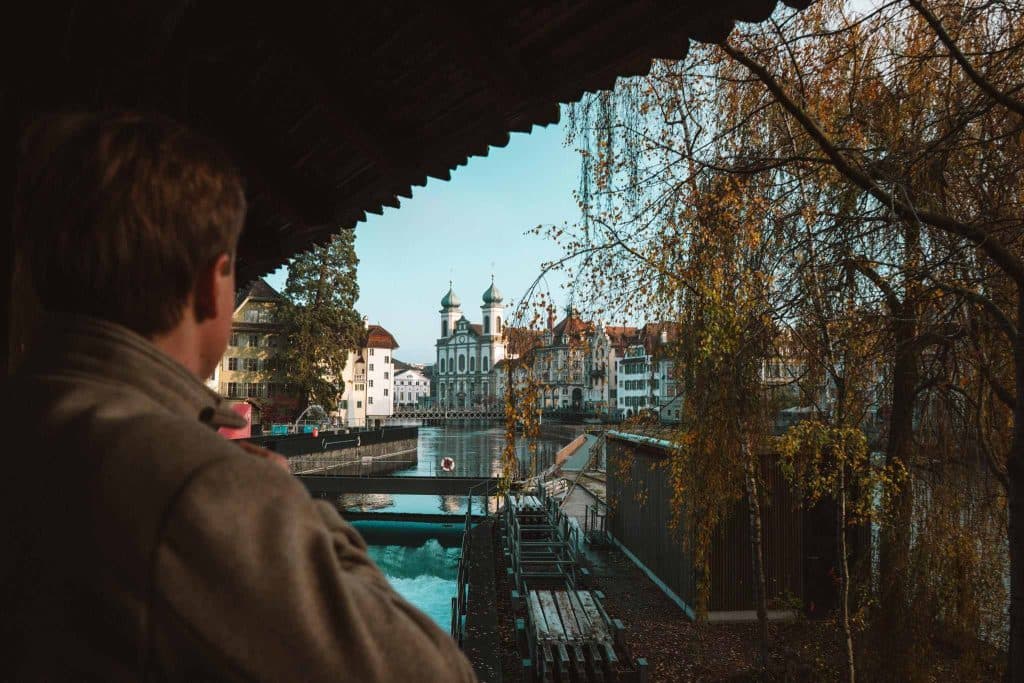 Een persoon staat onder een overdekte houten loopbrug die uitkijkt over een rustige rivier met historische gebouwen, een kerk met twee koepels en herfstbomen op de achtergrond.
