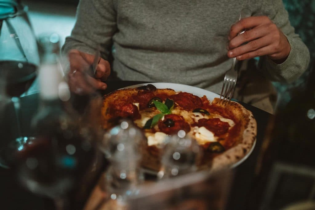 Een persoon in een grijze trui staat op het punt een pizza te eten met een vork en mes aan een schemerige tafel, met glazen wijn en water zichtbaar op de voorgrond.