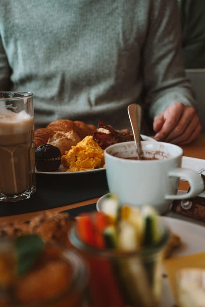 Een persoon zit aan een tafel met een bord roerei, spek, een croissant en een muffin, samen met een glas ijskoffie en een kop warme chocolademelk. Verse groenten zijn vaag op de voorgrond.