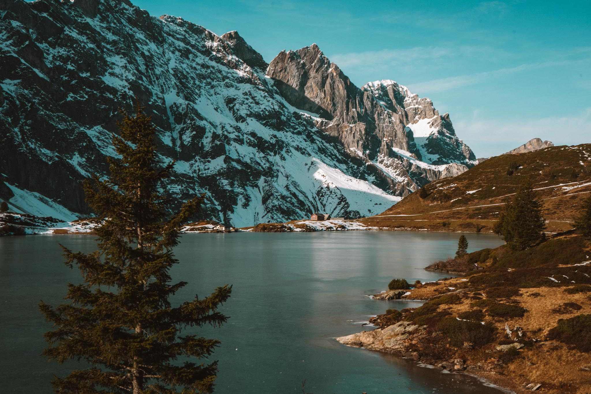 Een sereen bergmeer met helder water, omringd door groenblijvende bomen. Op de achtergrond rijzen besneeuwde rotsbergen op onder een blauwe hemel, waardoor een vredig, natuurlijk landschap ontstaat.