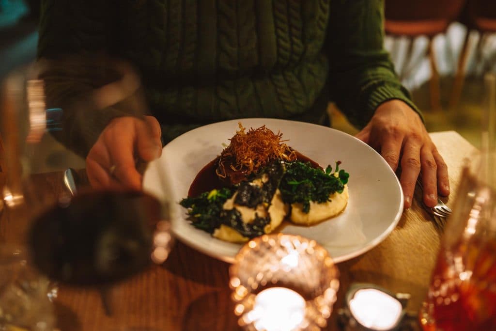 Een persoon in een groene trui zit aan een tafel en houdt een vork vast. Hij staat op het punt om een maaltijd te eten van aardappelpuree, groente, saus en knapperig garnituur, met glazen en kaarsen op de voorgrond.