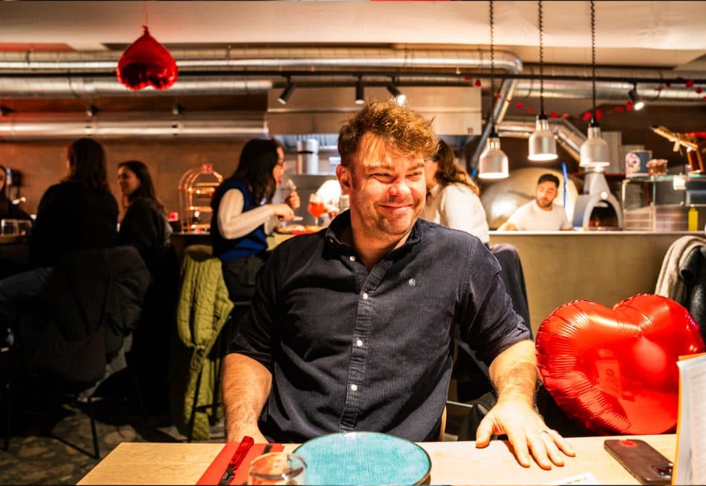 Een lachende man zit aan een restauranttafel in Leuven met een rode hartvormige ballon naast hem. Andere eters en rode hartversieringen zijn zichtbaar in de levendige, warm verlichte achtergrond - een viering van de liefde in een van de beste restaurants van de stad voor 2026.