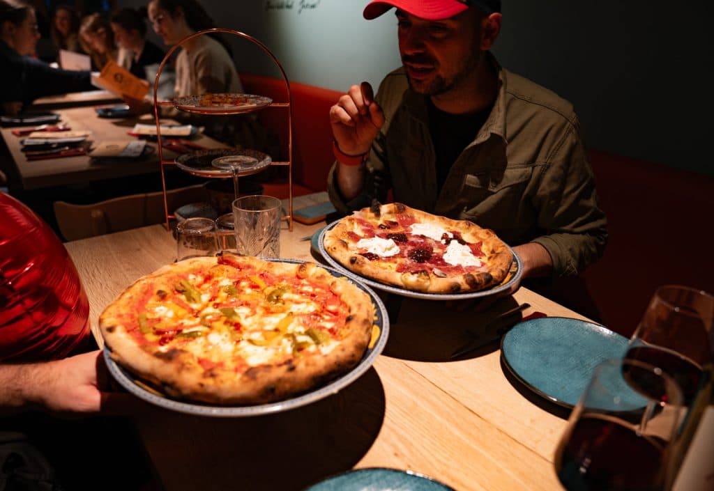 Twee mensen zitten aan een restauranttafel in Leuven met twee grote pizza's, borden en glazen rode wijn. De ene geniet van een groentepizza terwijl de andere vlees en kaas neemt - de essentie van dineren in een van de beste restaurants in Lekker Leuven.