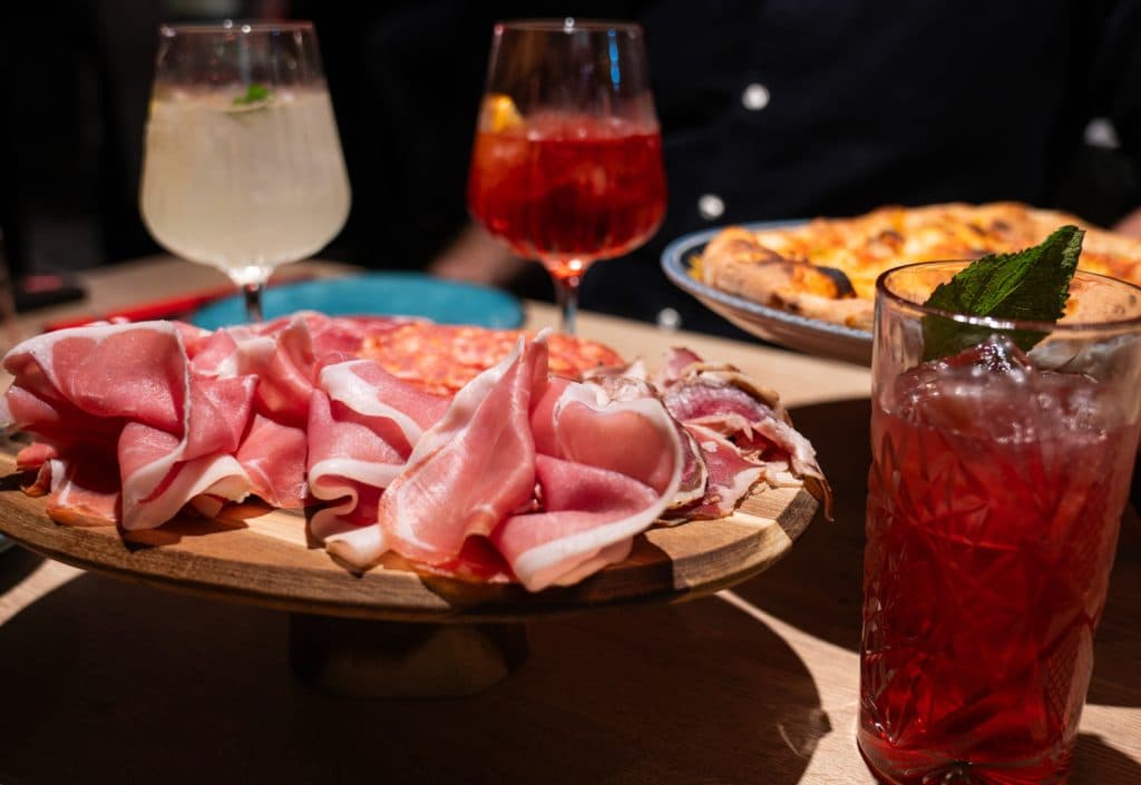 Een houten schotel met dungesneden prosciutto en salami staat op een tafel, naast cocktails en een bord pizza op de achtergrond - een beeld van de levendige sociale eetcultuur in veel Leuvense restaurants.
