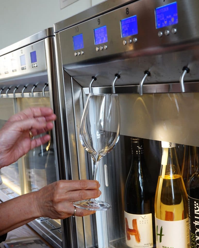 Een persoon houdt een wijnglas onder een dispenser, klaar om wijn in te schenken uit een geautomatiseerde machine met digitale displays.