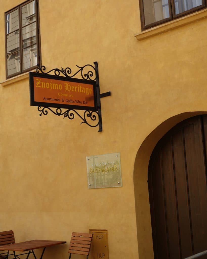 Een smeedijzeren bord met de tekst "Znojmo Heritage: Apartments & Gothic Wine Bar" hangt op een geel gepleisterd gebouw boven een grote gebogen houten deur.