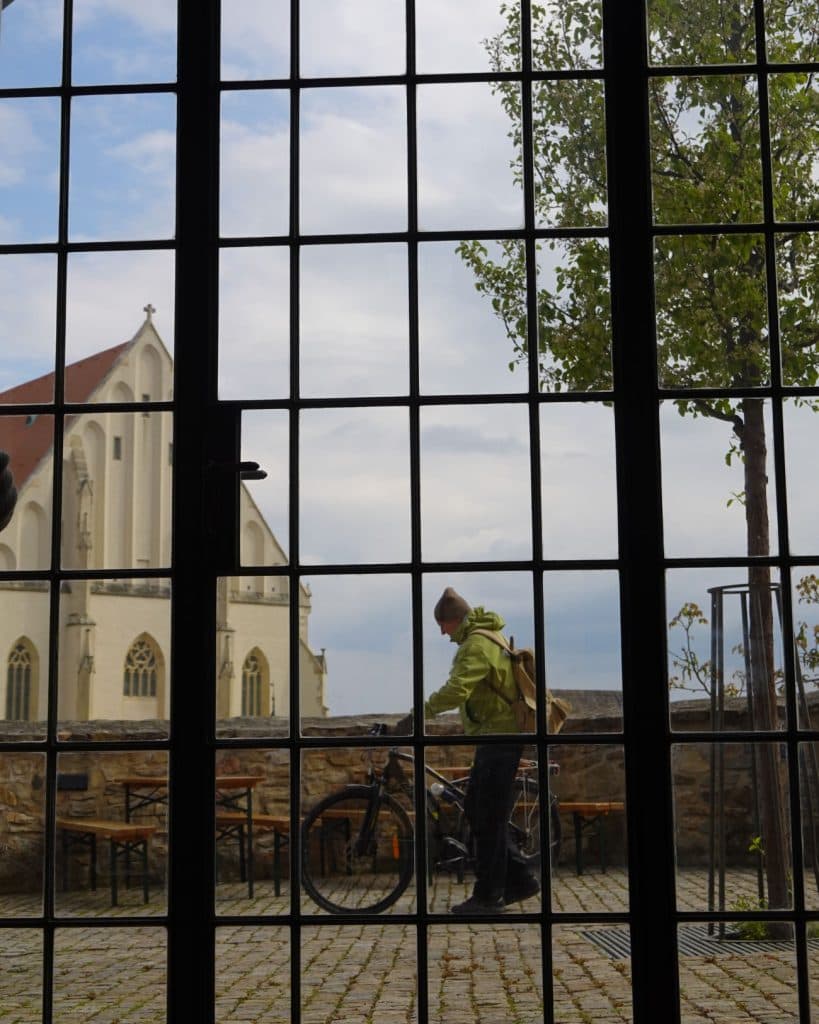 Een persoon in een groene jas staat naast een fiets op een geplaveide patio, gezien door een groot raam met een zwart rasterframe. Buiten wijzen lege stoelen en tafels op het nabijgelegen café Prins, met een kerk en een boom onder de bewolkte hemel.