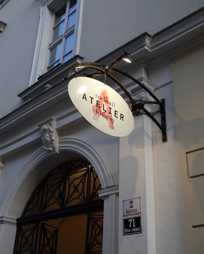Een wit ovaal bord met een rode afbeelding en zwarte tekst "Cocktail ATELIER & BISTRO" hangt boven een deuropening aan een smeedijzeren beugel in Brno, Tsjechië. Hieronder staat op een muurbord het adres: "2 Koblizna, 71 Brno-město.