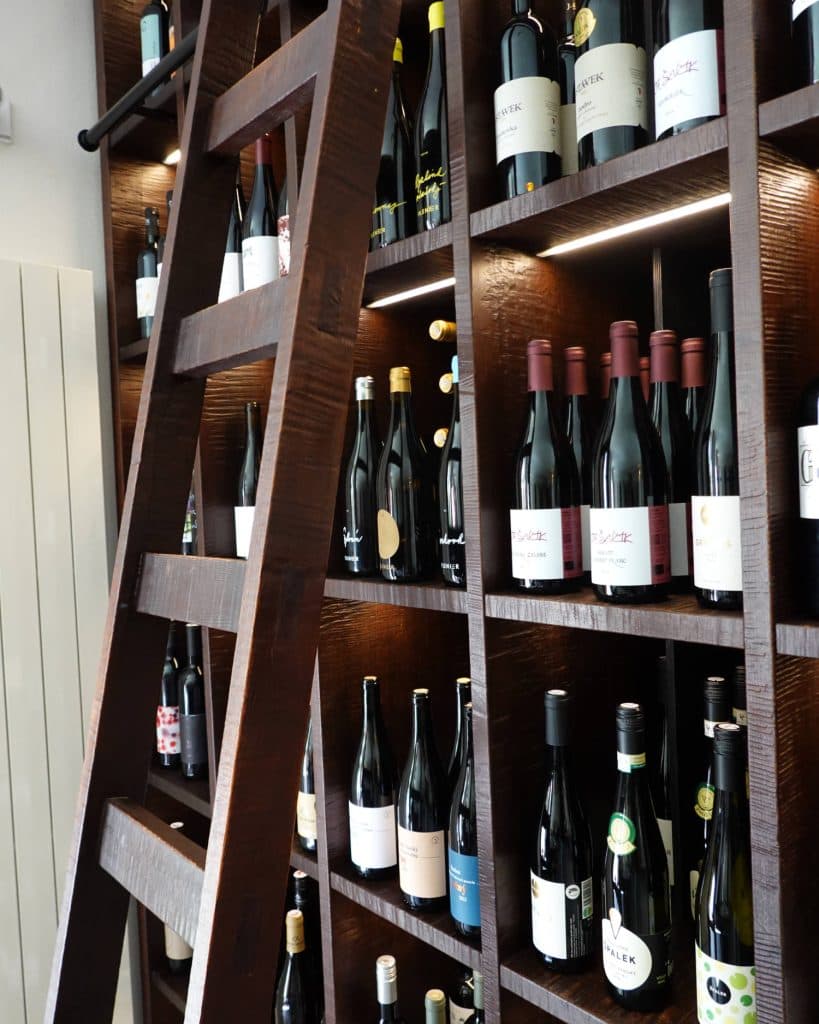 Een donkerhouten wijnrek gevuld met verschillende flessen wijn, sommige met een rode dop en andere met een witte, staat tegen een muur. Een bijpassende houten ladder leunt op het rek en doet denken aan een grote kelder in Tsjechië.