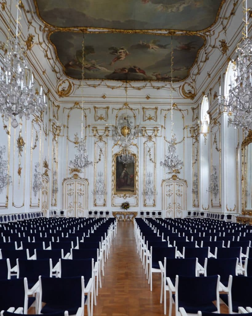 Sierlijke witte en gouden balzaal met hoog, uitvoerig versierd plafond met muurschilderingen, kristallen kroonluchters, hoge ramen en rijen marineblauwe stoelen - geschikt voor een prins of prinses van Tsjechië - met uitzicht op een centraal schilderij en voetstuk op de gepolijste houten vloer.
