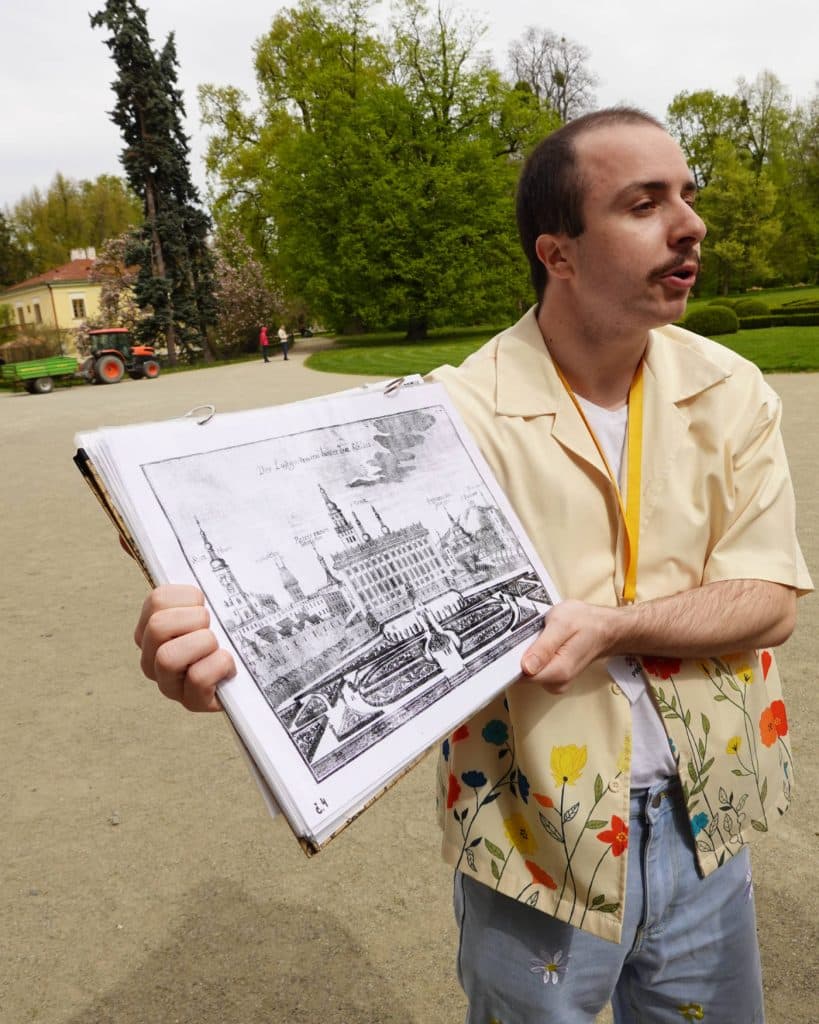 Een man met kort haar en een snor, die een geel overhemd met bloemenmotief en een lichtblauwe broek draagt, staat buiten in Tsjechië en toont een grote zwart-wit illustratie van een historisch gebouw. Op de achtergrond zijn groene bomen en een tractor te zien.