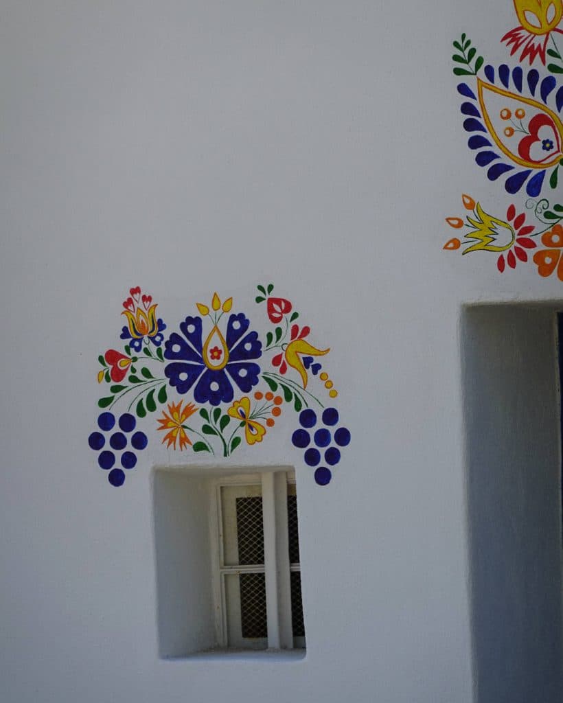 Een klein, rechthoekig raam met witte spijlen zit in een witte muur in Tsjechië. Boven en naast het raam vormen kleurrijke, florale volkskunstmotieven in blauw, geel, rood, groen en oranje een prinsesachtig symmetrisch patroon.