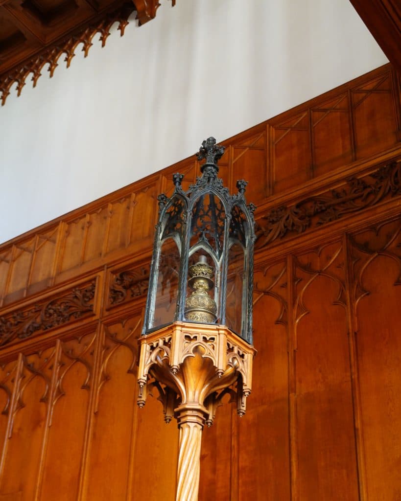 Een decoratieve houten paal met een sierlijke gotische glazen en metalen kast bovenop, waarin een gouden voorwerp is opgeborgen, staat te midden van ingewikkeld houtsnijwerk in een historisch interieur in Tsjechië. Het licht valt op de lambrisering van een prinses.