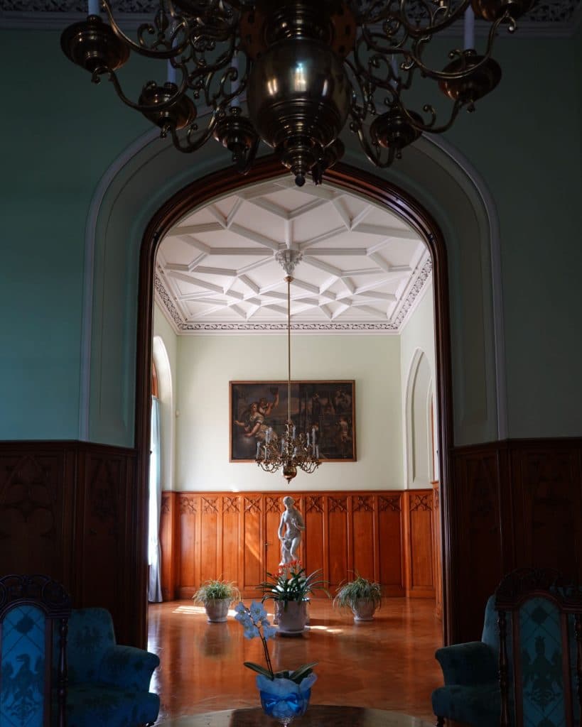 Een sierlijke kamer met groene muren in sprookjeskasteel, een grote gewelfde houten deuropening en een kroonluchter. Een beeldhouwwerk staat op de houten vloer, onder een andere kroonluchter, met blauwe stoelen en een orchidee op de voorgrond, als een tafereel dat past bij een prinses.